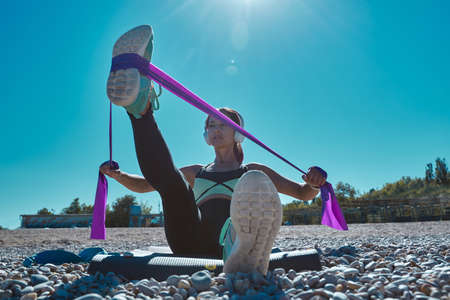 Fit woman stretching leg with rubber bands. Person train on yoga mat on beach. Active lifestyle and...