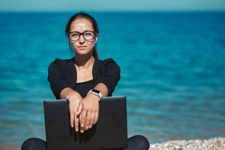 Clever woman sitting on beach with laptop. Cute seascape on backdrop. Travel time and freelance job idea, copy spaceの写真素材
