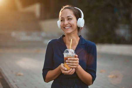Cheerful laughing female holding cup of drink. Woman wearing white headset outdoors. Take break and...