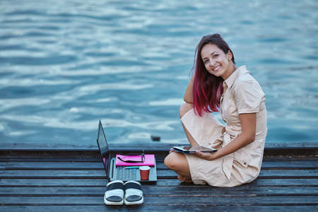 Smiling girl studying on pier near water. Laptop, notebook and cup of coffee. Distance learning and...