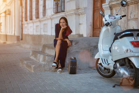 Lady sit on step with suitcase. White motorbike on right side and building on backdrop. Travel time...