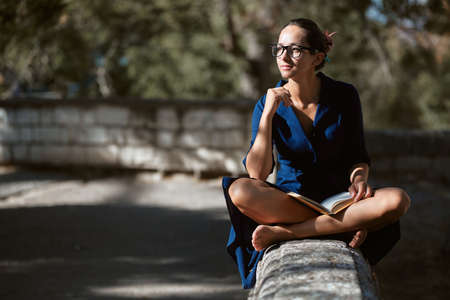 Pensive young woman in glasses looking away sitting in park. Street style, read book and city walk concept, copy spaceの写真素材