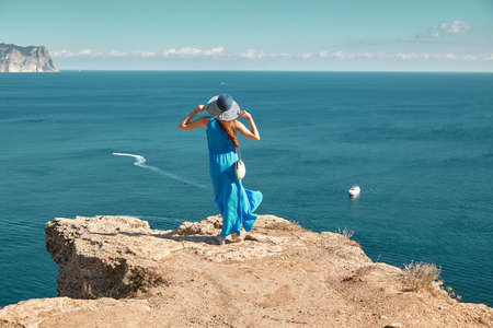 Woman in blue dress and hat enjoying ocean view from mountain. Floating yacht in sea. Travel time and new ground ideaの写真素材