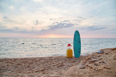 Travelling set, surfboard and suitcase. Sunset over sea, sandy coastline. Summer vacation and surfing idea. Copy spaceの写真素材
