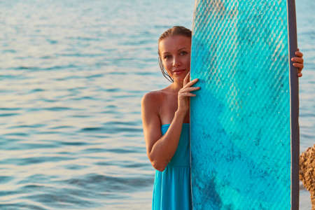 Smiling woman posing with surfboard an seashore. Happy girl surfer in blue dress. Summertime and time to adventure ideaの写真素材