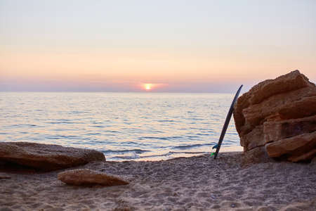 Nice sunset view on beach. Surfboard on rock and calm sea. Summer time, surfing sport and time to rest idea, copy spaceの写真素材