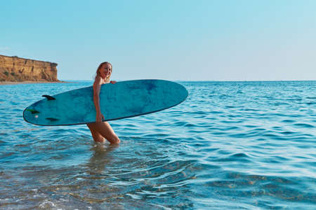 Happy girl walk in ocean with surfboard. Calm sea view on summer day. Idea of surf sport, beach day...