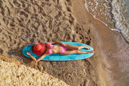Woman lay on surfboard on sandy beach cover face with hat. Summer time, surf and travel time, desire of adventure ideaの写真素材
