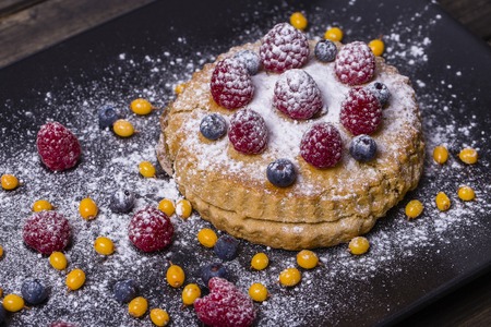 Cake with raspberries, blueberries, sea buckthorn sprinkled with powdered sugar on a black plate, close upの写真素材
