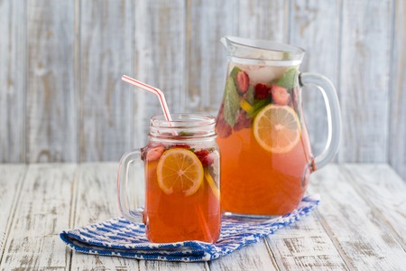 Dietary detox drink with lemon juice, red strawberry and mint leaves in clear water with ice on wooden table. Pink lemonade close up in mason jar mugの写真素材