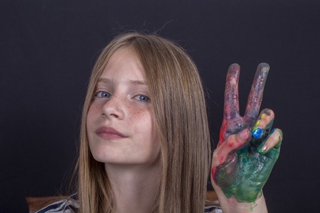 Beautiful blond young girl with freckles and hands painted in colorful paints indoors on black...