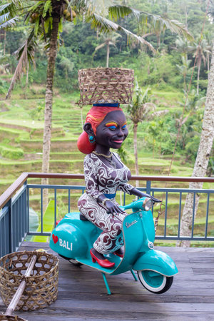 BALI, INDONESIA - MARCH 25, 2018 : Figure of a dark-skinned woman sits on a motorcycle Vespa with a wicker basket on her head in tropical cafe near rice terraces of Tegallalang in Bali, Indonesiaのeditorial素材