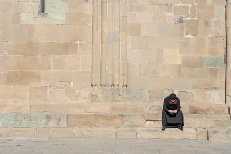 MTSKHETA, GEORGIA - OCTOBER 27, 2018 : lonely old monk in black clothes sits on the stairs on a sunny day near the old orthodox cathedral in historical town Mtskheta near Tbilisi, Georgiaのeditorial素材