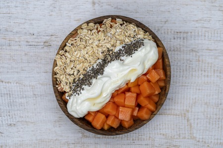 Slices of ripe sweet papaya fruit with oat flakes, chia seeds and white yogurt on coconut bowl on white wooden background, close up . The concept of healthy eatingの写真素材