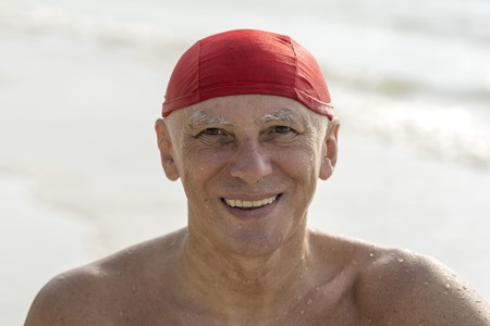 Happy elderly man in a red swimming hat on the beach near the sea waterの写真素材