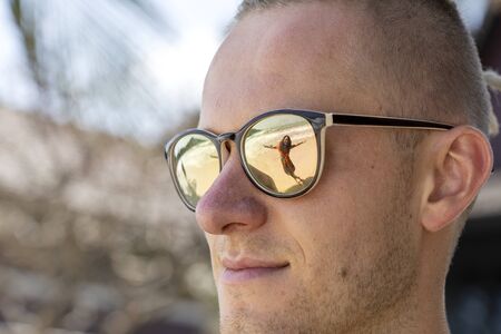 Portrait of a young man in sunglasses in which the silhouette of a girl is reflected on the tropical beach near sea, close upの写真素材
