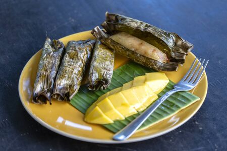 Thai style dessert, yellow mango with banana sticky rice in palm leaves. Yellow mango and sticky rice is popular traditional dessert of Thailand. Close upの写真素材