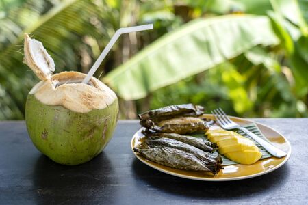 Thai style dessert, yellow mango with banana sticky rice in palm leaves and green coconut. Yellow mango and sticky rice is popular traditional dessert of Thailand. Close upの写真素材