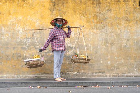 Hoi An, Vietnam - march 16, 2020 : Vietnamese woman in a straw hat with a basket of fruits on a street market in the old city against the background of the old yellow wall in Hoi An, Vietnamのeditorial素材