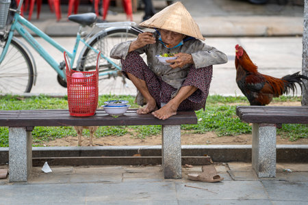 Hoi An, Vietnam - march 16, 2020 : A Vietnamese old woman in a straw hat sits on a bench and eats food on a street in the old city in Hoi An, Vietnamのeditorial素材