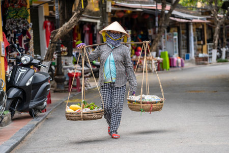Hoi An, Vietnam - march 16, 2020 : Vietnamese woman in a straw hat with a basket of fruits on a street market in the old city in Hoi An, Vietnamのeditorial素材
