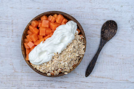 Slices of ripe sweet papaya fruit with oat flakes and white yogurt on coconut bowl on white wooden...