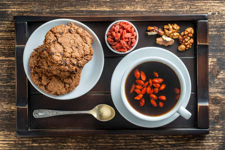 White cup of fresh morning goji coffee with cookies on wooden tray on the table, top view, close up. Black hot coffee with whole red goji berriesの写真素材