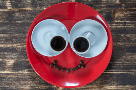 Red plate along with two white cups of coffee and a smile from coffee beans on a wooden table. Close up, top viewの写真素材