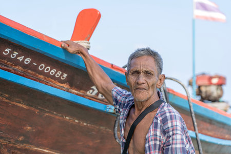 KOH PHANGAN, THAILAND - FEBRUARY 07, 2019: Thai old fisherman is standing near his wooden boat near the shore on the island Koh Phangan, Thailandのeditorial素材