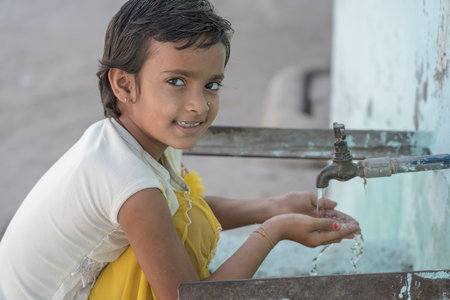 RISHIKESH, INDIA - NOVEMBER 06, 2018: Portrait of thirsty Indian girl drinks water from the outdoor tap on the street in Rishikesh, Indiaのeditorial素材