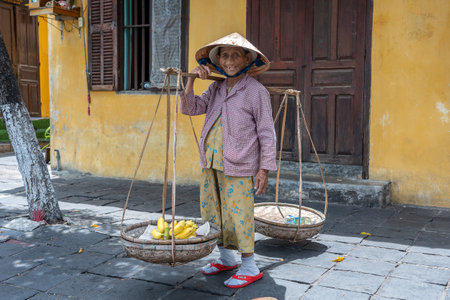 Hoi An, Vietnam - July 01, 2020: Vietnamese woman in a straw hat with a basket of fruits on a street market in the old city against the background of the old yellow wall in Hoi An, Vietnamのeditorial素材