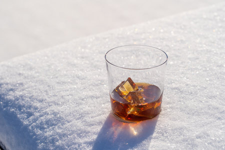 Glass of whiskey with ice on a bed of snow and white background, close up. Concept of christmas winter morningの写真素材