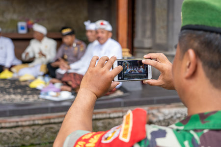 Ubud, island Bali, Indonesia - March 06, 2019: Indonesian military man photographs a group of priests on a cell phone during a holiday in island Bali, Indonesiaのeditorial素材