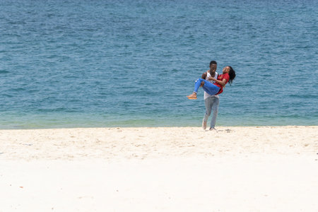 Zanzibar, Tanzania - November 03, 2019: A young African guy holds a girl in his arms on a sandy beach near sea on the island of Zanzibar, Tanzania, East Africaのeditorial素材