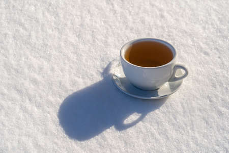 White cup of hot tea on a bed of snow and white background, close up. Concept of christmas winter morningの写真素材