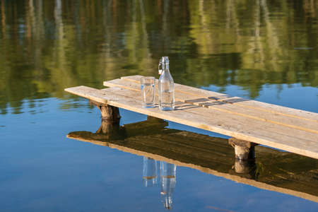 Glass and bottle with drinking water on a wooden pier in the morning near the lake and forest in spring time, close up. Nature and travel conceptの写真素材
