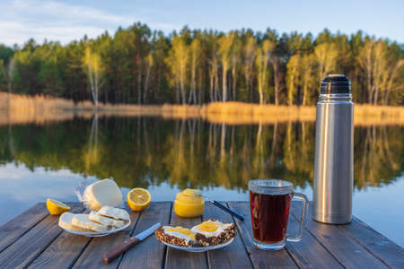 Breakfast with hot tea, cheese sandwich and honey on wooden table in the morning next to the lake...