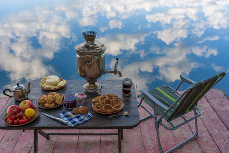 Vintage metal tea samovar with white smoke and food on the table near the calm water lake in green...