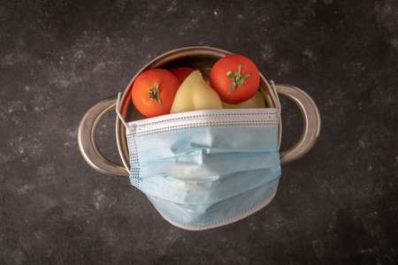 Funny face with protective mask on a saucepan of vegetables on a black background, close up. Face made of saucepan, tomato and pepper with medicine protective maskの写真素材