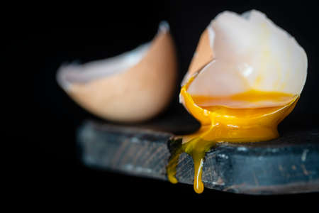 Broken egg shell with dripping yolk on wooden cutting board on black background, close up, macro