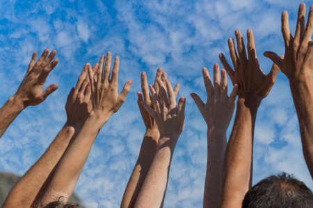 Group of people raise hands in air across blue sky with white clouds, close up