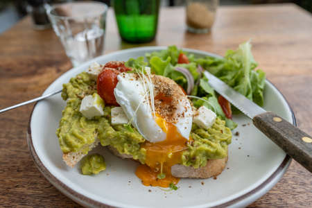 Sourdough toast, poached eggs, avocado pulp and fresh vegetables on plate in cafe, close up,...