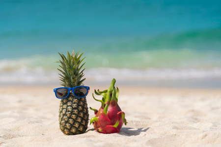 Ripe attractive pineapple in sunglasses and pink dragon fruit on the sand tropical beach against turquoise sea water, Thailand. summer vacation conceptの写真素材