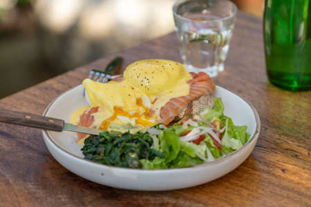 Sourdough toast, poached eggs, salmon, hollandaise, sauteed and fresh vegetables on plate in cafe, close upの写真素材