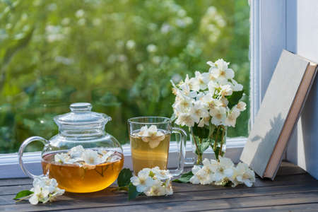 Delicious hot herbal tea on the windowsill at home at summer day near garden and beautiful bouquet of jasmine flowers, close up. Hot jasmine tea in a glass teapot and cupの写真素材