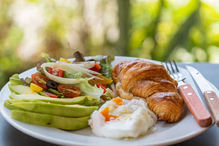 Croissant sandwich with poached egg, tomato and avocado on plate in cafe, close up