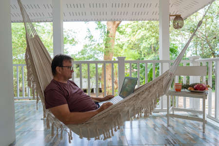 Grown man rests and working a laptop in a hammock on a terrace home near tropical garden in island Koh Phangan, Thailand, close upの写真素材