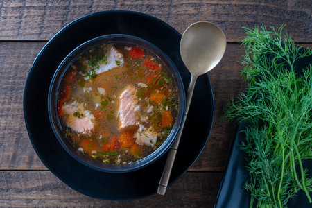 Fresh salmon fish soup with carrots, potatoes and red peppers in a black plate on wooden table, close up. Tasty dinner consists of a fish soup with salmon. top viewの写真素材
