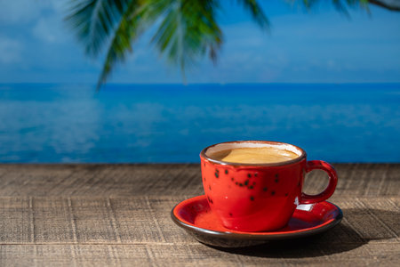 Red mug of hot black coffee on wooden table with sea water, coconut palm tree and blue sky background on sunny summer day in tropical beach cafe, close up, copy spaceの写真素材
