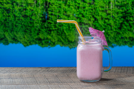 Healthy cold strawberry smoothie in glass mug on wooden table with the lake water and forest background on a sunny summer day, close upの写真素材